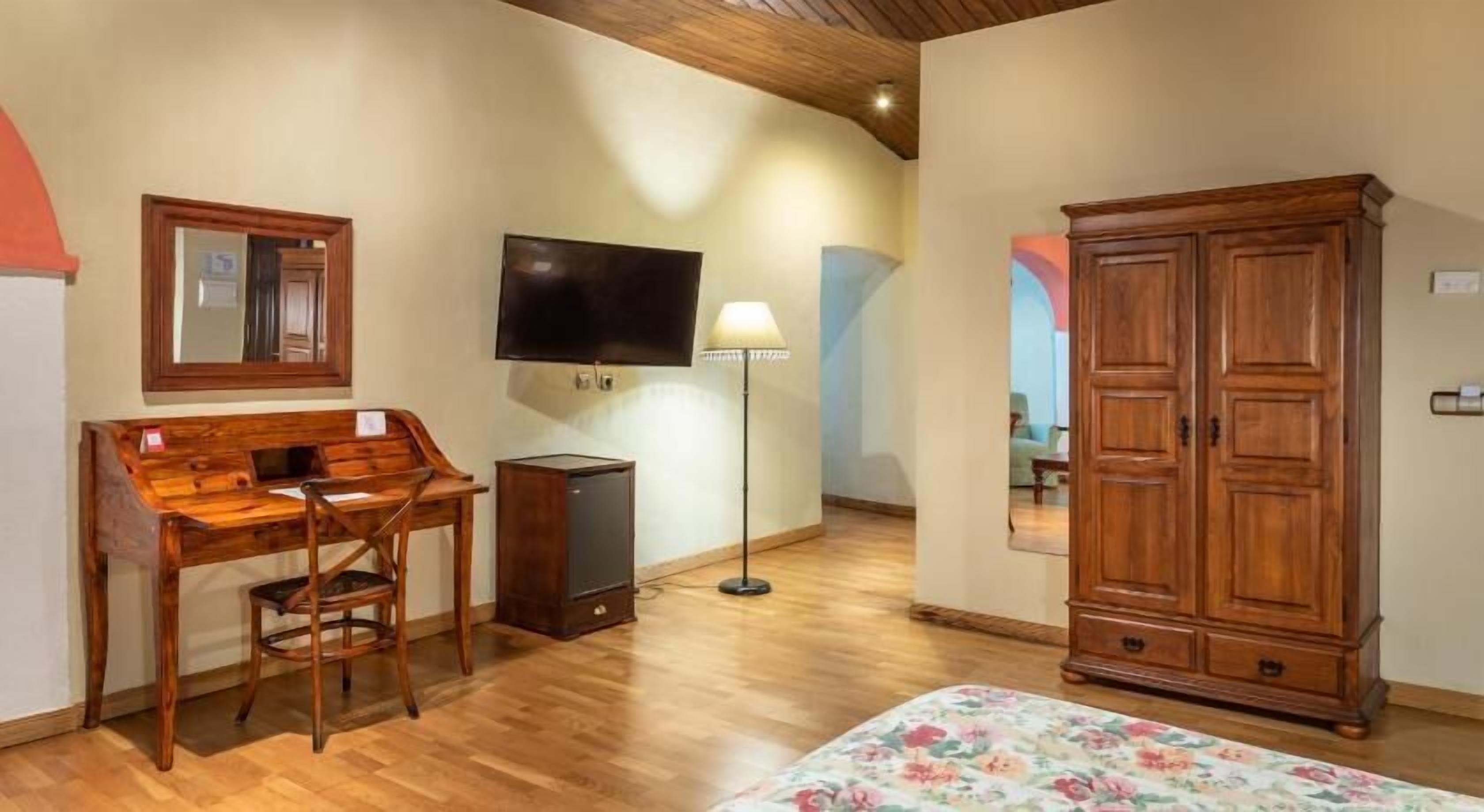 A room with a wooden desk, a television, a floor lamp, a wardrobe, and a bed with a floral bedspread.