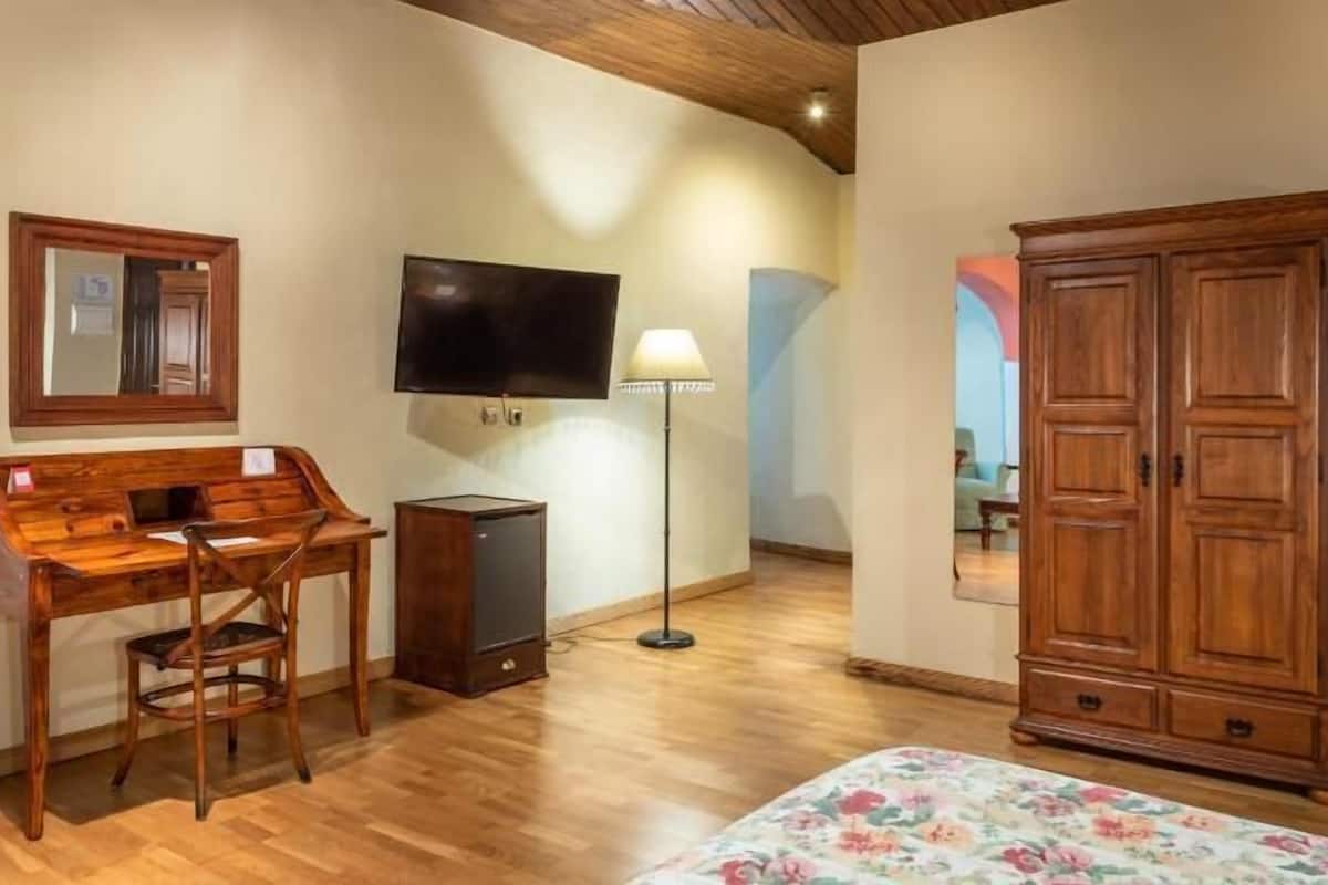 A room with a wooden desk, a television, a floor lamp, a wardrobe, and a bed with a floral bedspread.