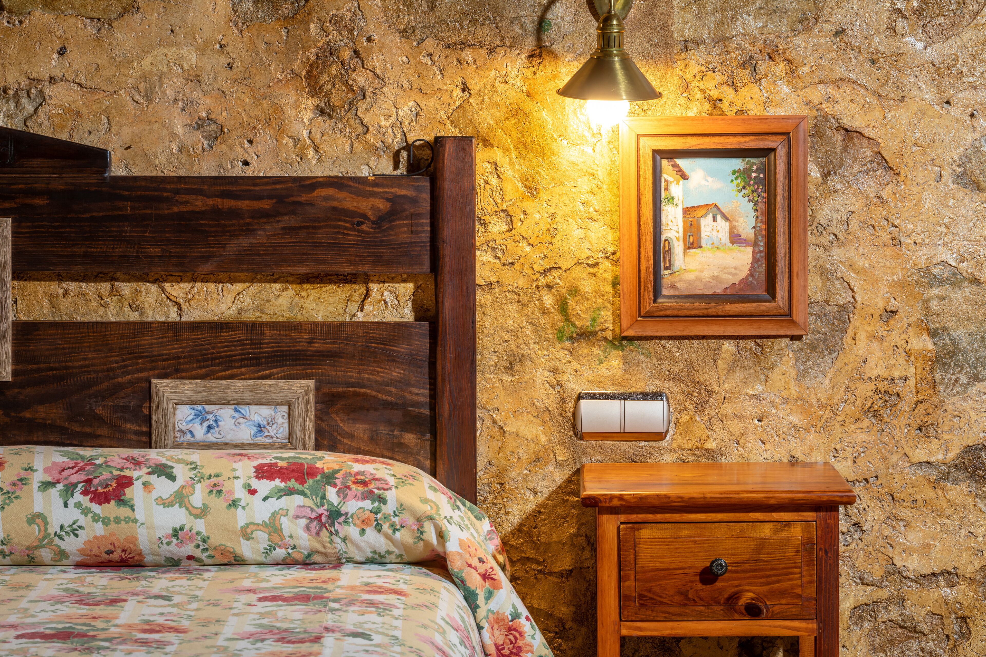 Ein Schlafzimmer mit Holz-Kopfteil, floralem Bettbezug, einem Nachttisch mit Schublade, einem gerahmten Bild an der Wand und einer Wand mit Steinoptik.