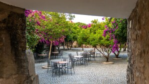 Un cafè a l'aire lliure amb taules i cadires disposades a un pati de pedra, envoltat d'arbres amb flors roses.