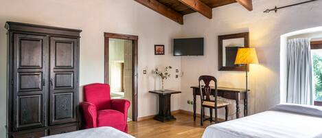 A room with two beds, a red chair, a wooden cabinet, a table with a lamp, and a TV on the wall.