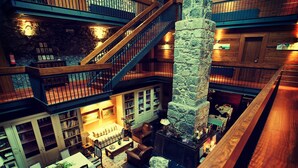 A multi-level hotel lobby with a stone fireplace, wooden staircase, and a dining area with a red sofa and tables.