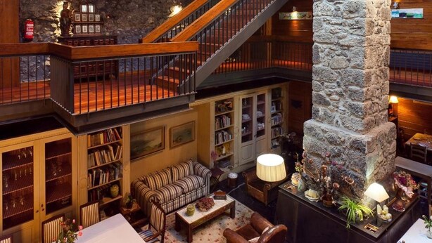 A multi-level interior with a stone fireplace, wooden staircase, and a cozy seating area with a red sofa and patterned armchairs.
