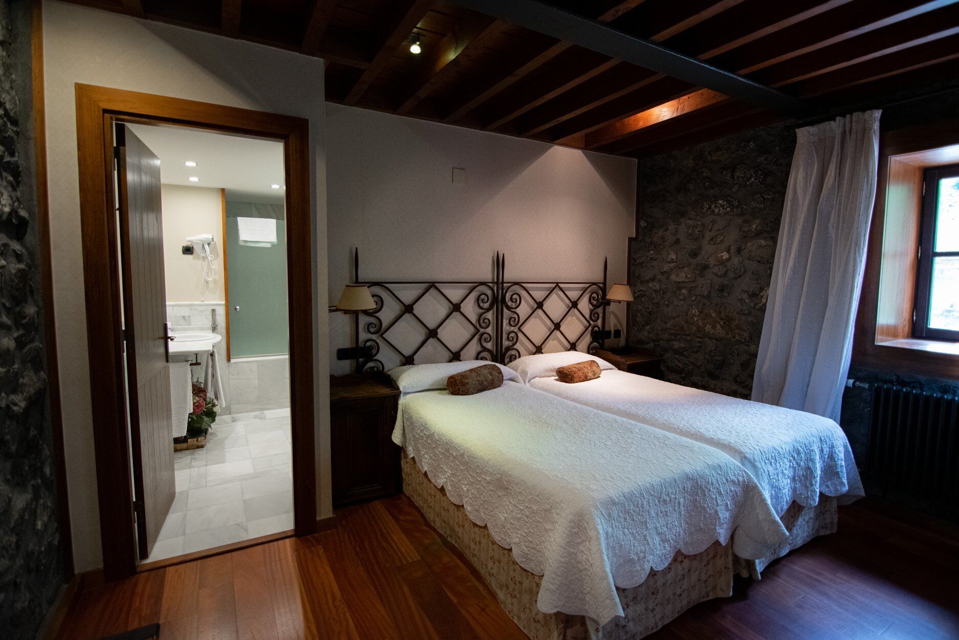A bedroom with a wooden ceiling, a stone wall, a bed with white bedding, a nightstand, and a window with curtains.