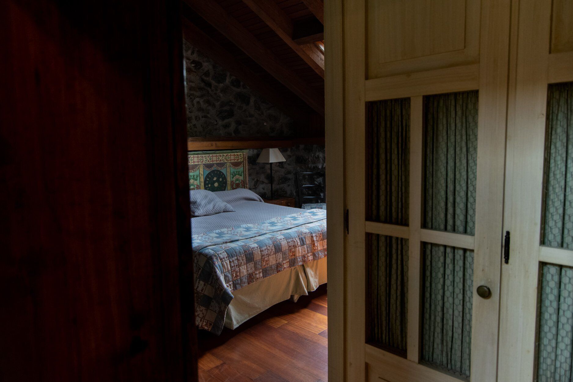 A bedroom with a bed, a nightstand, and a lamp. The room has wooden flooring and a stone wall.
