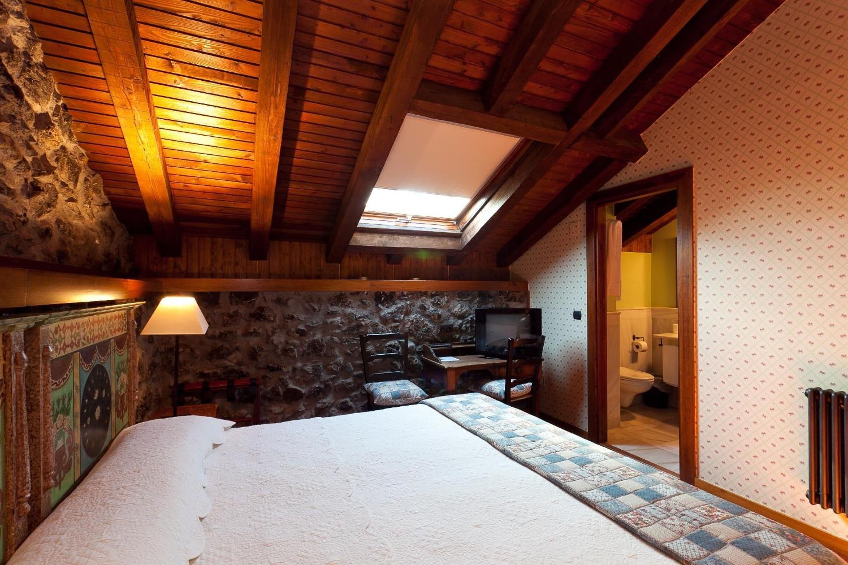 Un dormitorio acogedor con techo de madera, pared de piedra, cama con colcha, escritorio con silla, televisor y baño visible a través de una puerta abierta.