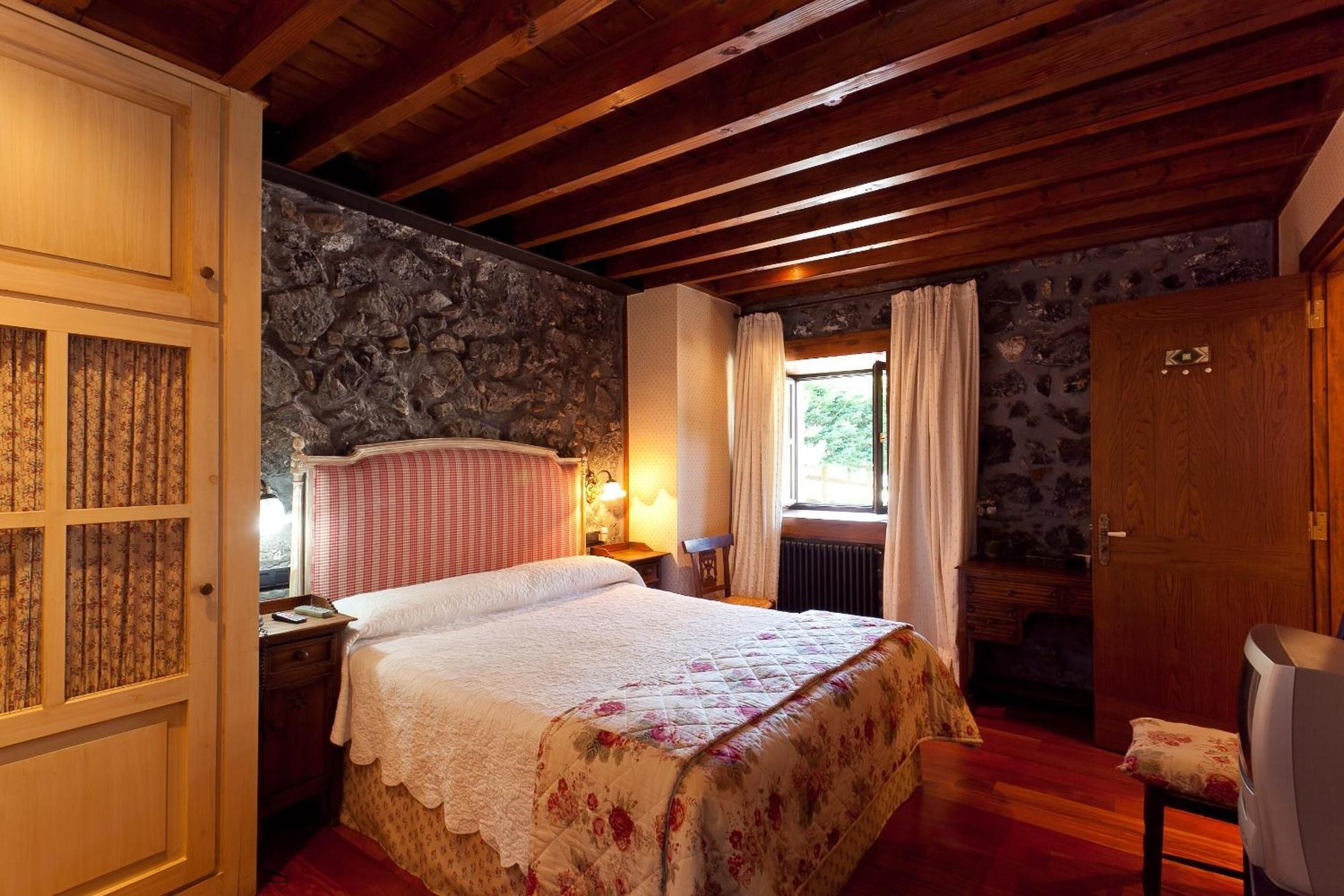 Un dormitorio acogedor con techo de madera, pared de piedra, cama con colcha estampada, mesita de noche, cómoda y ventana con cortinas.