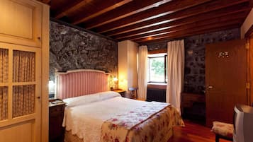 Un dormitorio acogedor con techo de madera, pared de piedra, cama con colcha estampada, mesita de noche, cómoda y ventana con cortinas.