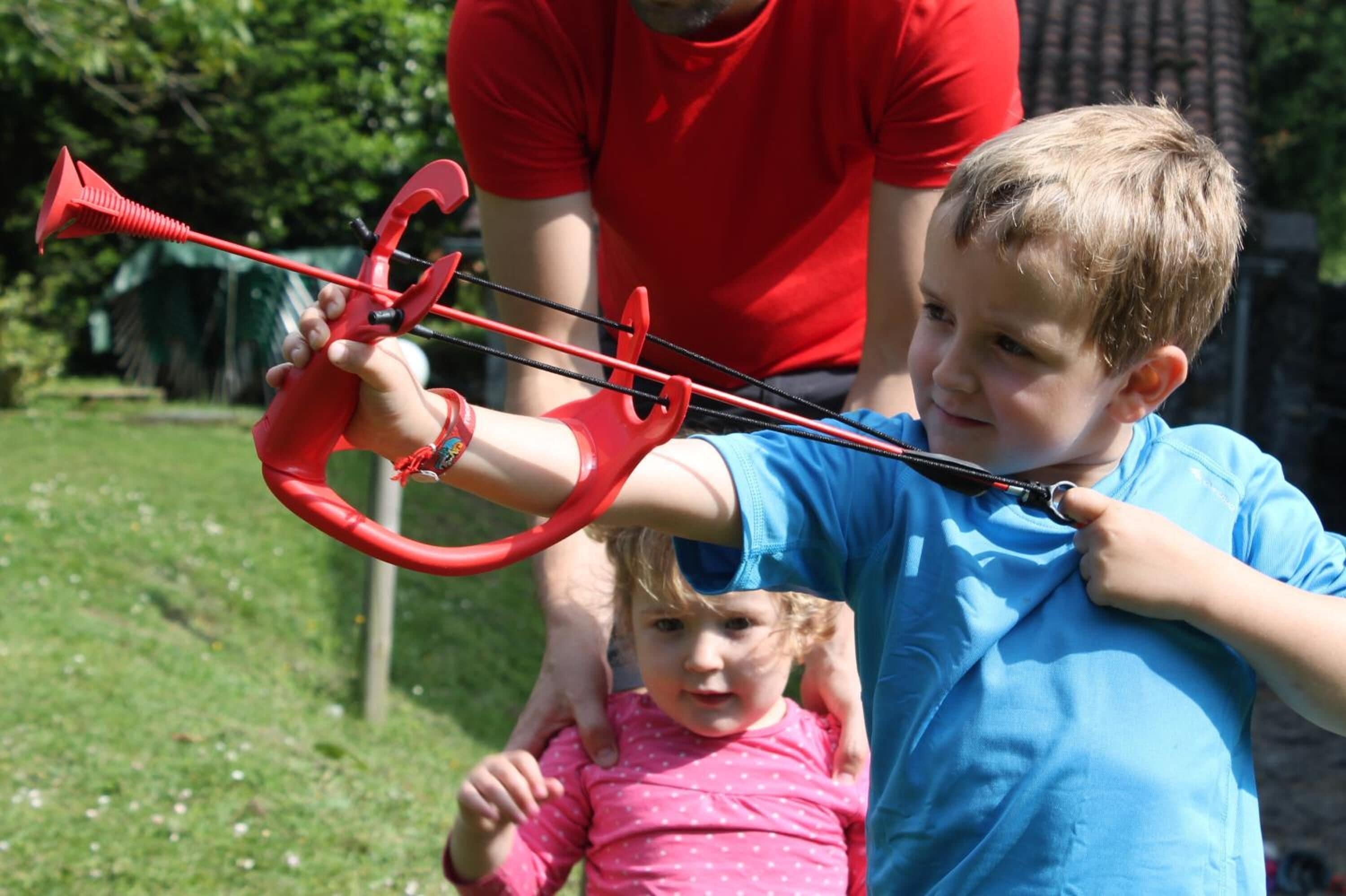 A child holding a red toy bow and arrow, with an adult in a red shirt standing behind.