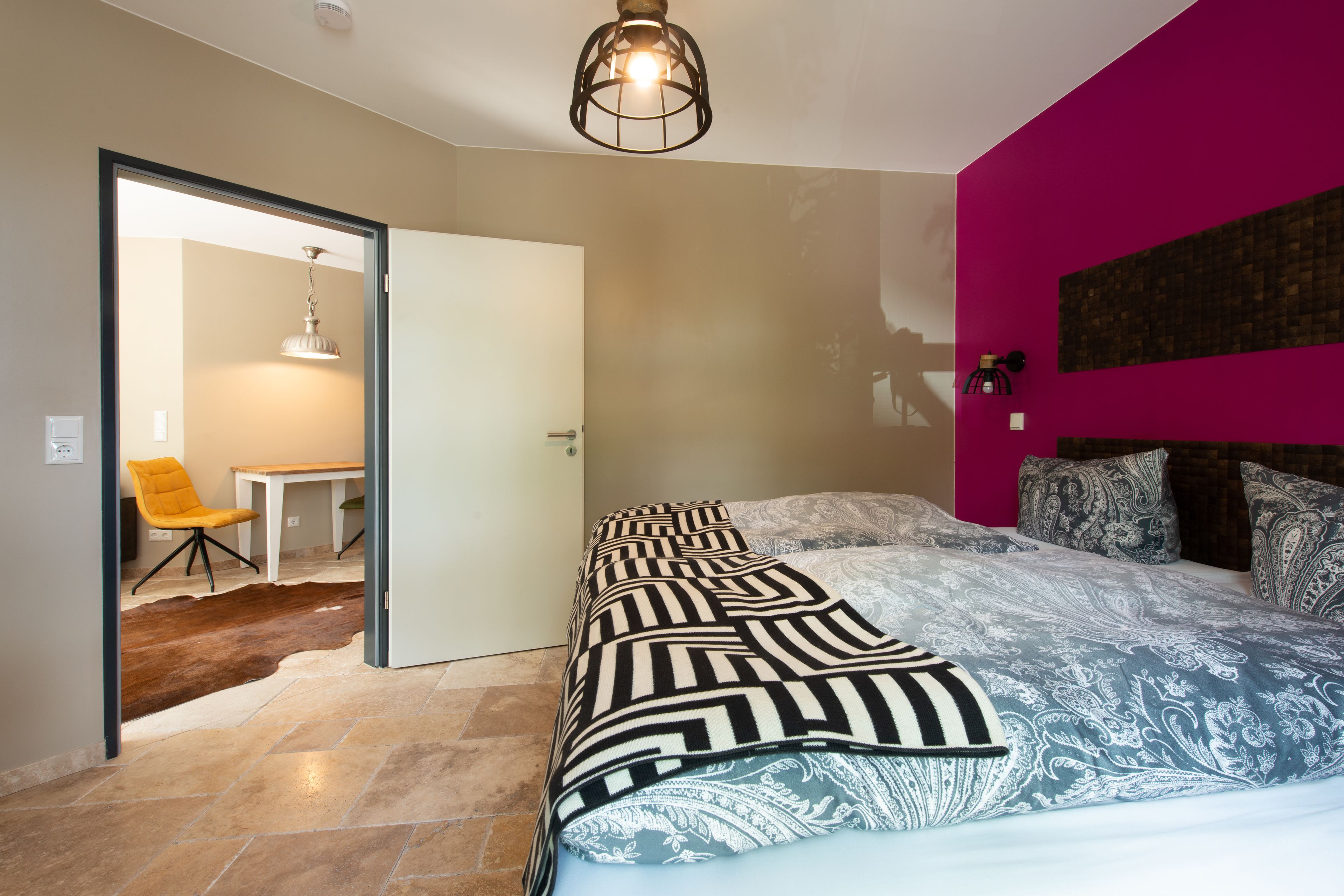 A bedroom with a bed, a chair, a table, and a door leading to another room.