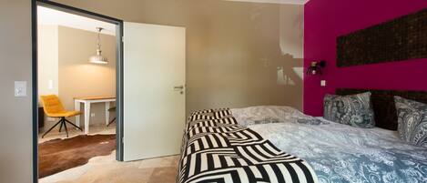 A bedroom with a bed, a chair, a table, and a door leading to another room.