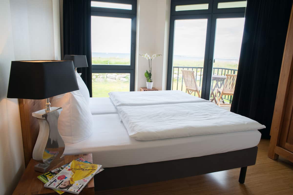 Une chambre d’hôtel avec un lit, une table de chevet avec une lampe, un magazine et une vue sur la plage grâce à de grandes fenêtres.