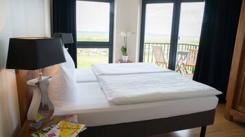 Habitación de hotel con cama, mesita de noche con lámpara, una revista y vista a la playa a través de amplias ventanas.