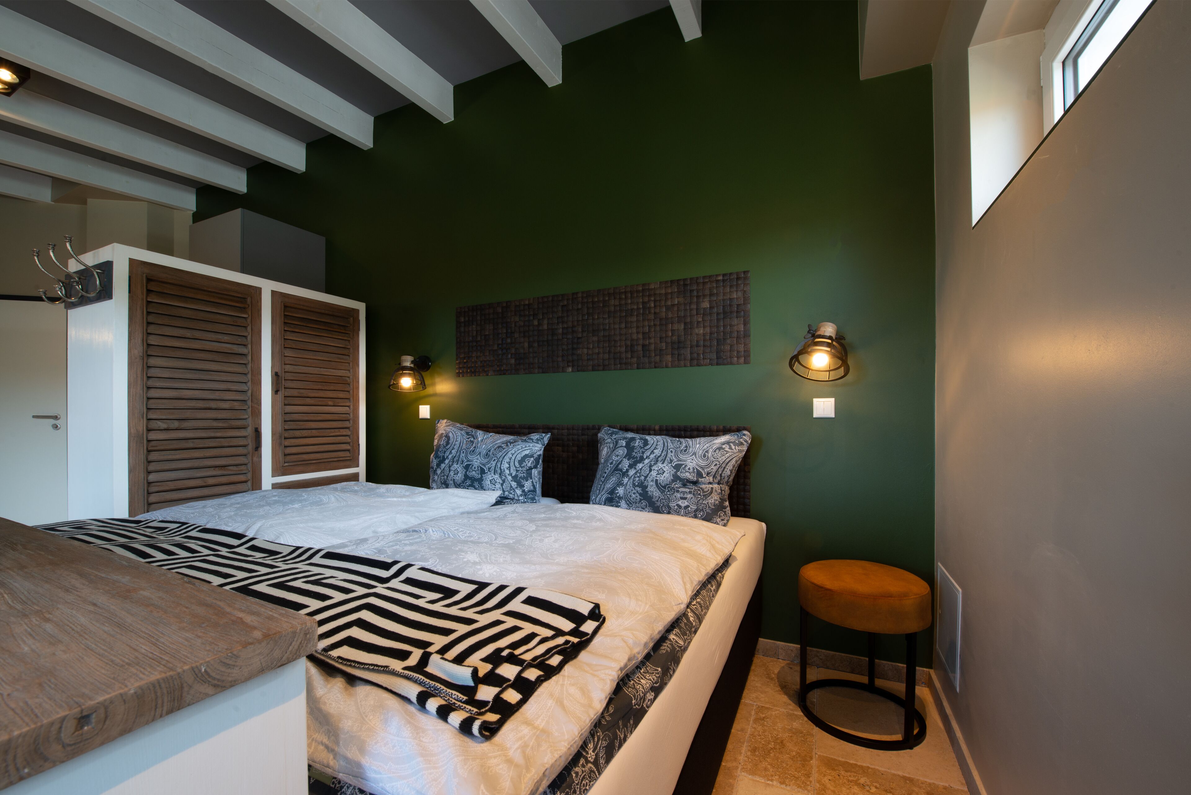 Une chambre à coucher avec un lit, un banc en bois, une lampe fixée au mur et une fenêtre.