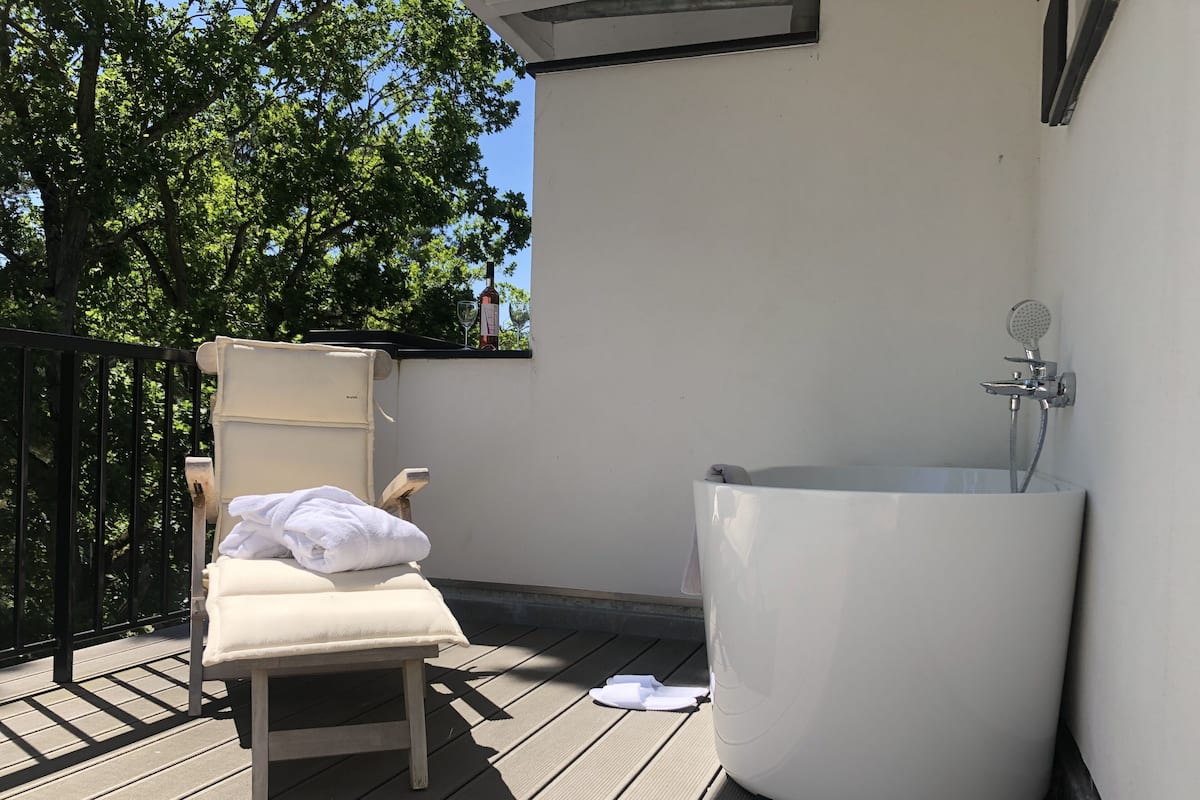 Un balcon avec une baignoire blanche, un fauteuil de couleur beige et un verre de vin posé sur la balustrade.