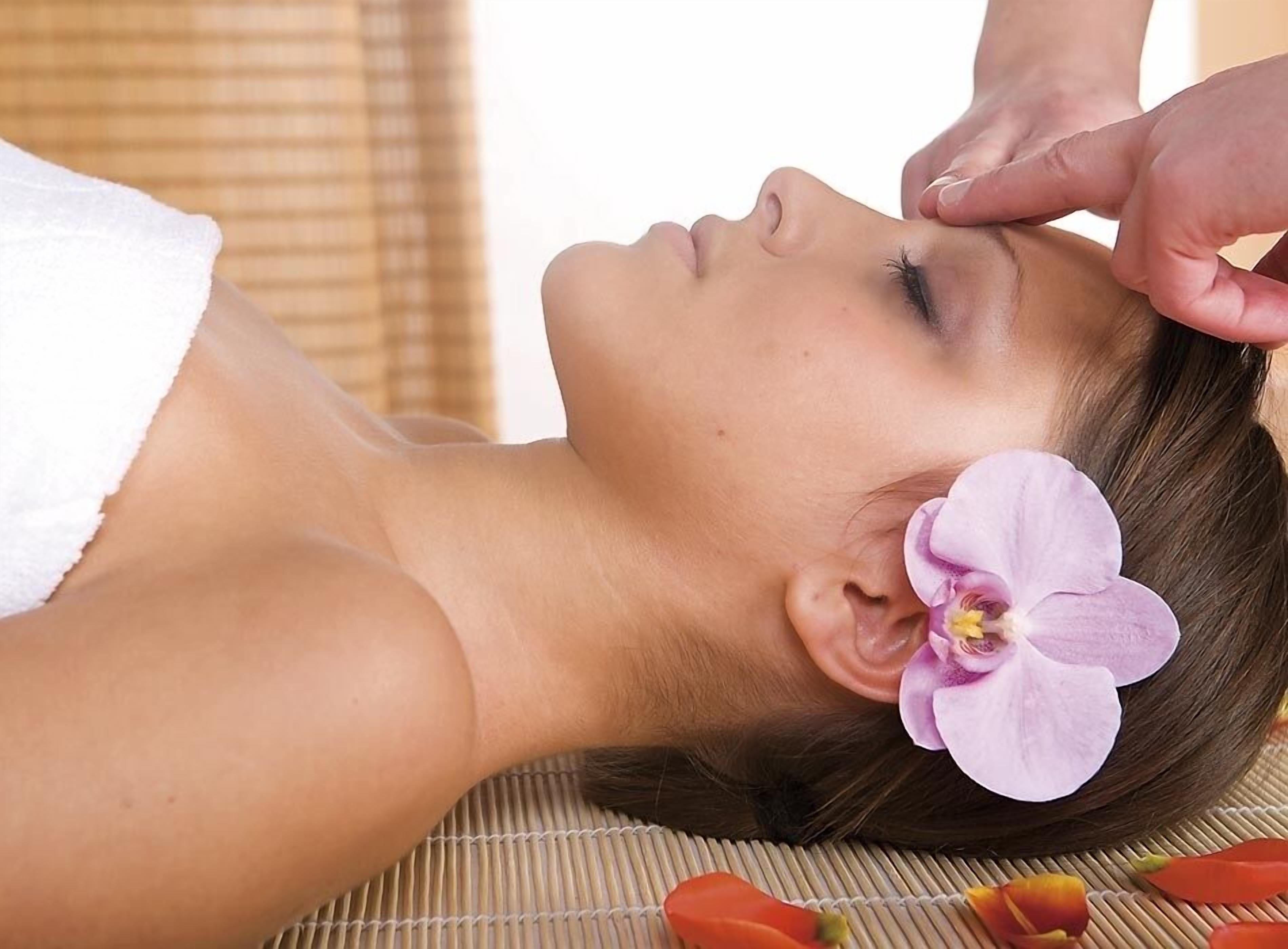 A person lying down with a towel on their shoulder, a flower in their hair, and a massage being performed on their neck.