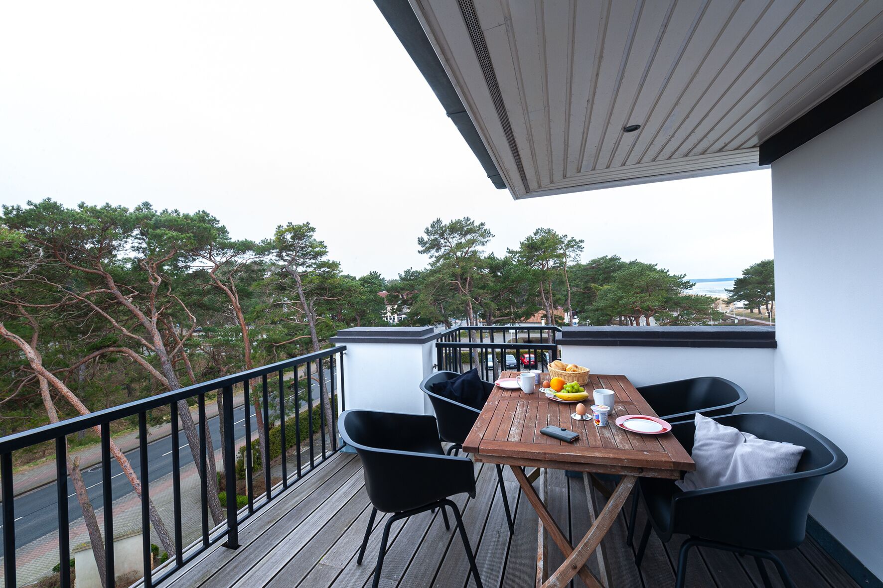Una terraza de madera con una mesa dispuesta para el desayuno, rodeada de sillas negras, con vista a una carretera y árboles.