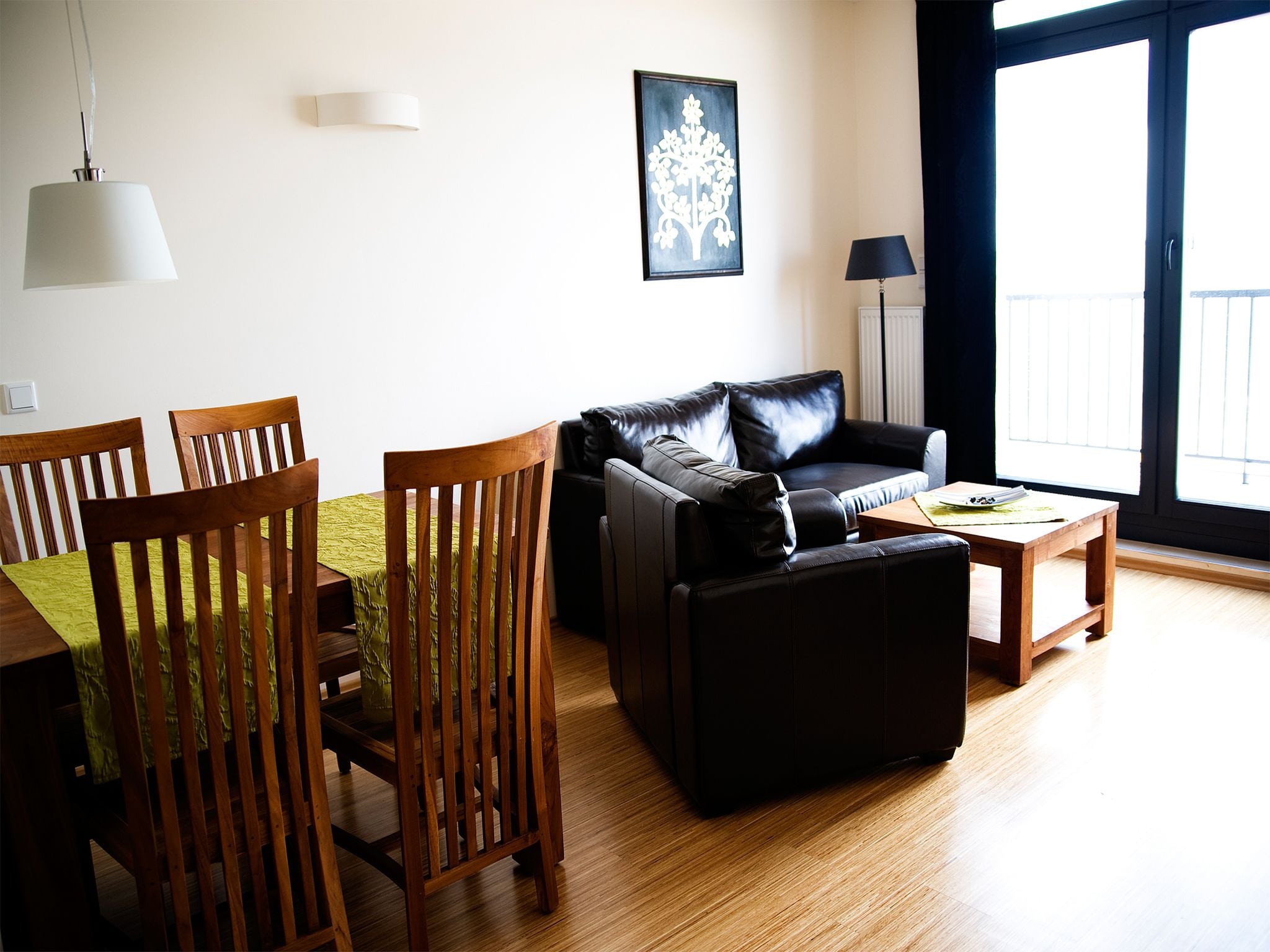 A living room with a leather sofa, wooden chairs, a dining table with a green tablecloth, a coffee table, and a large window with a view.