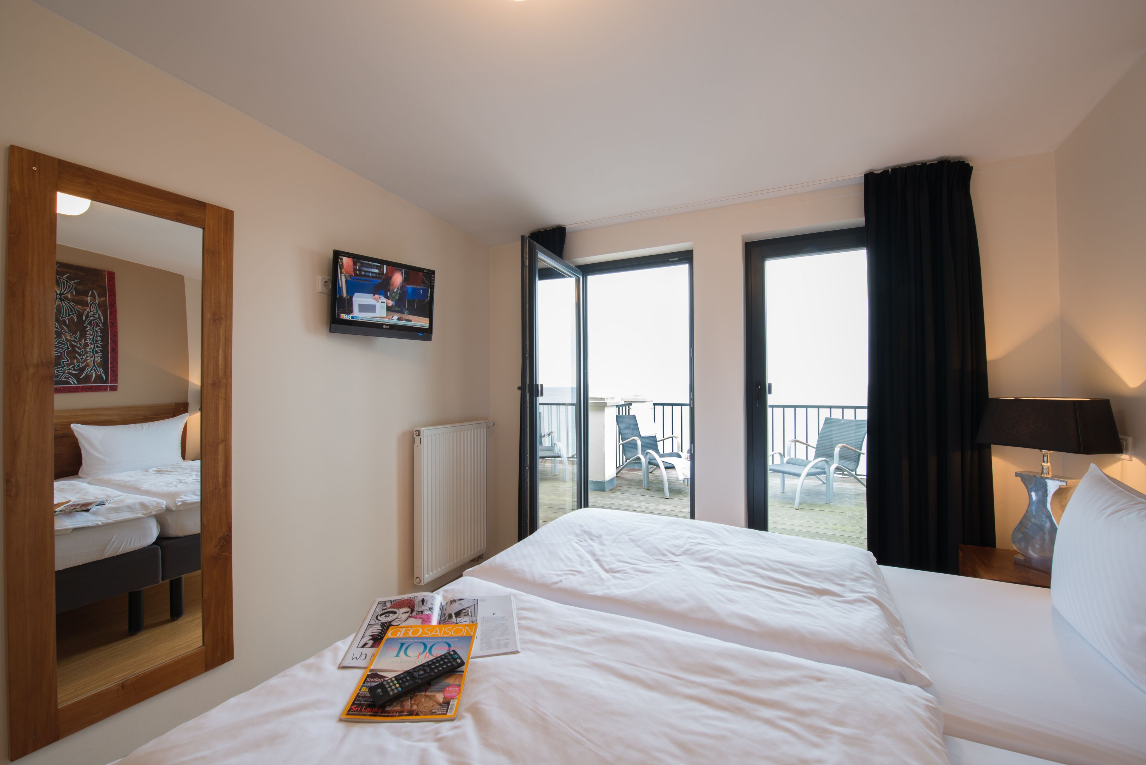 A hotel room with a large bed, a television on the wall, a balcony with chairs, and a window with curtains.
