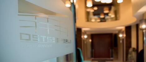 Een hotelgang met een glazen wand waarop het logo “OBTSEER PERLE” staat afgebeeld, en een receptie met een trap naar een verdieping erboven.