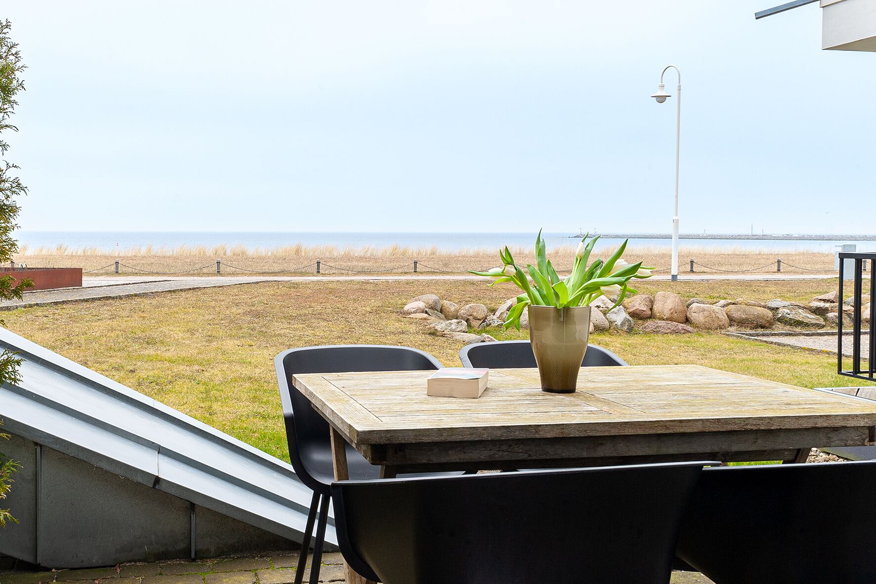 A table with a vase of green plants, black chairs, and a view of the beach and ocean.