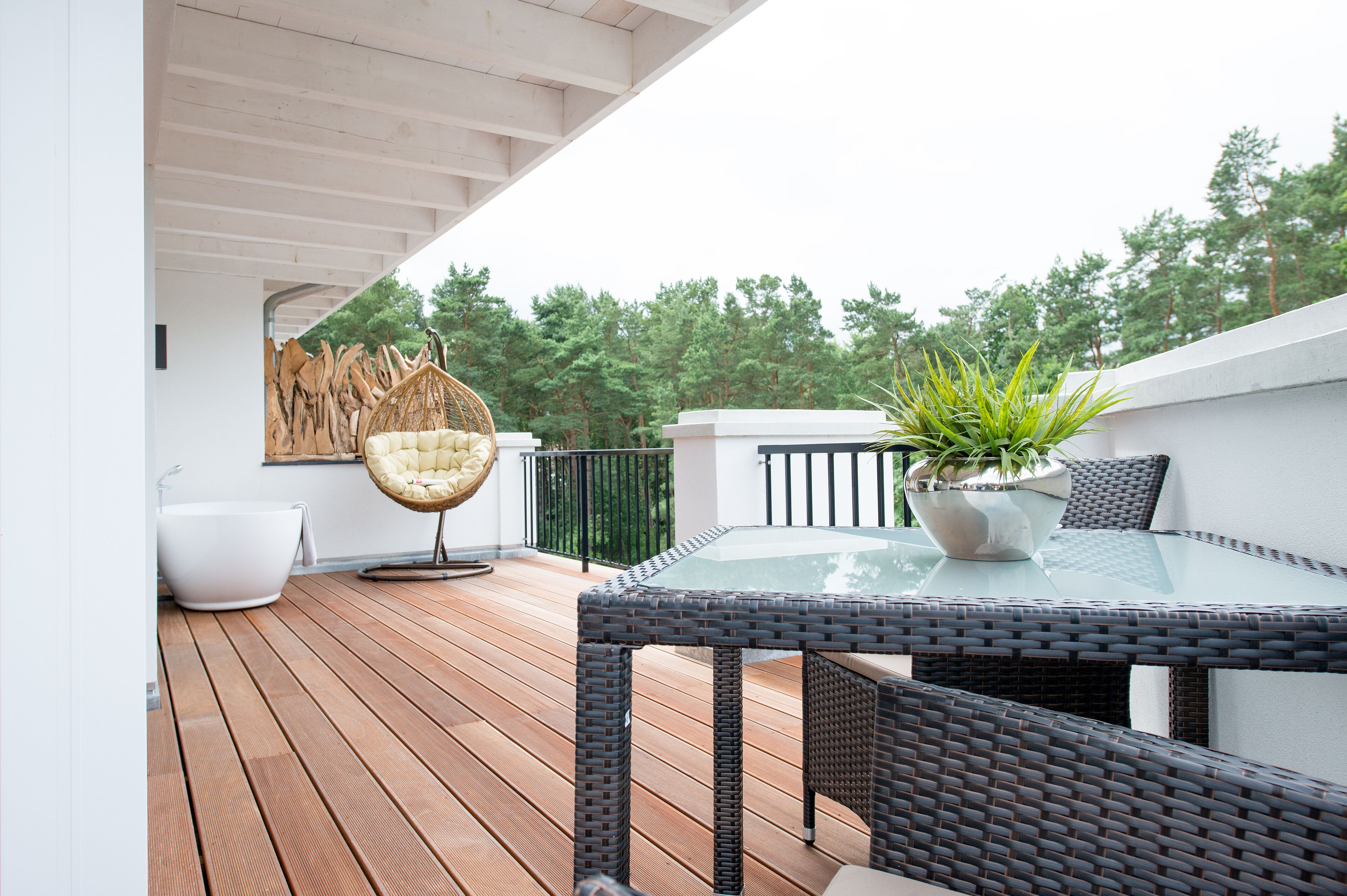 A wooden deck with a glass-top table, wicker chairs, a white bathtub, and a hanging chair.