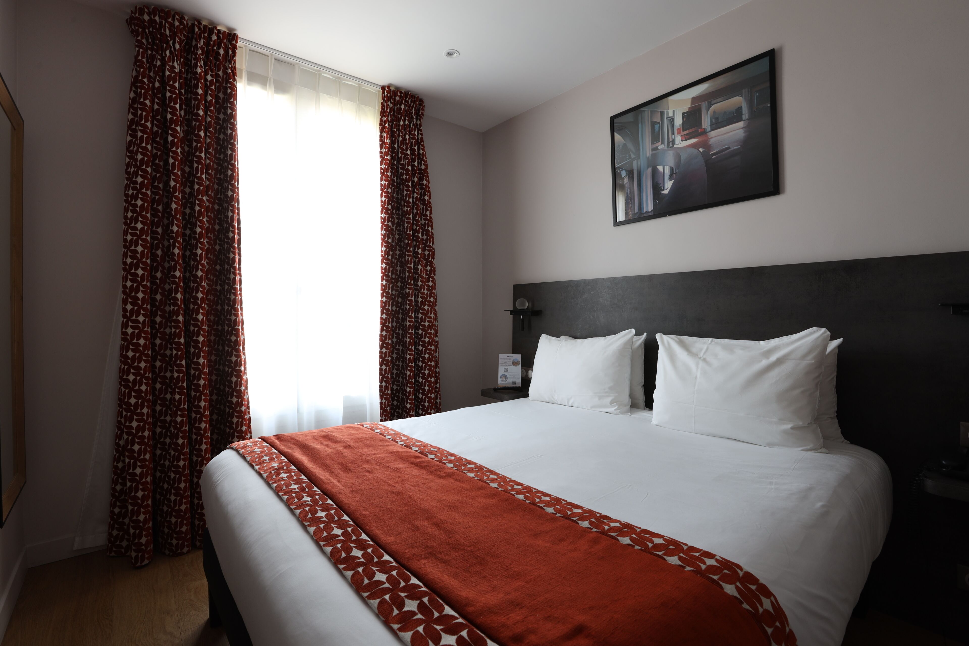 A hotel room with a bed, curtains, a nightstand, a wall-mounted TV, and a painting.