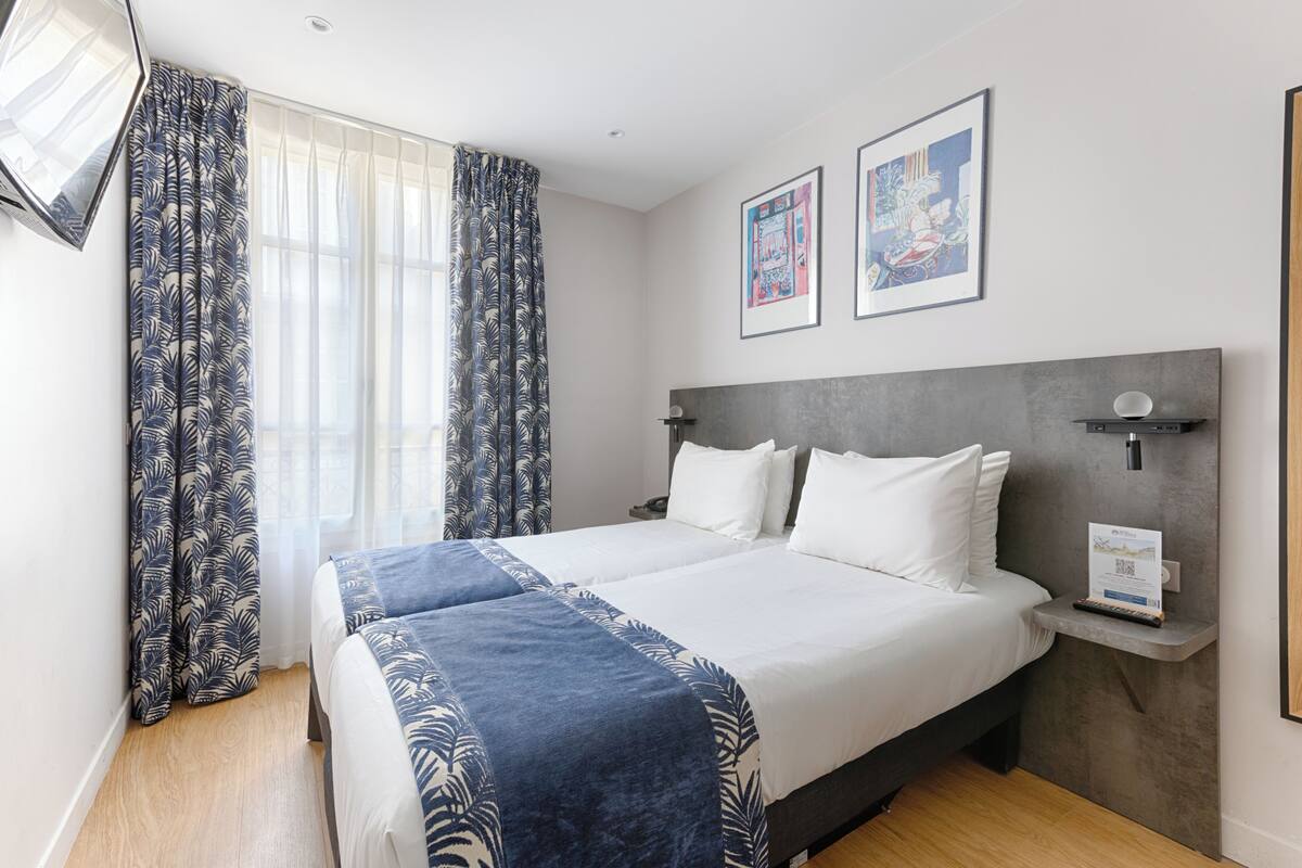 A hotel room with a double bed, a bedside table, a wall-mounted shelf, and a window with curtains.