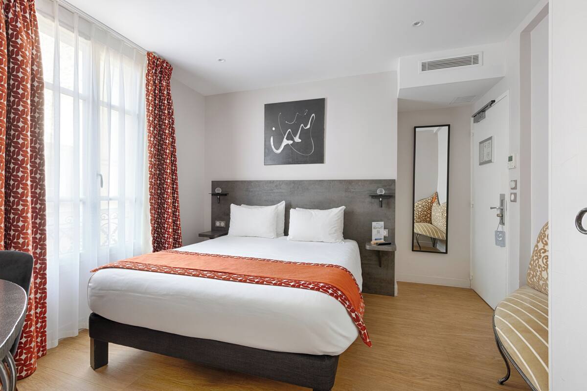 A hotel room with a bed, a desk, a chair, a mirror, and a door.