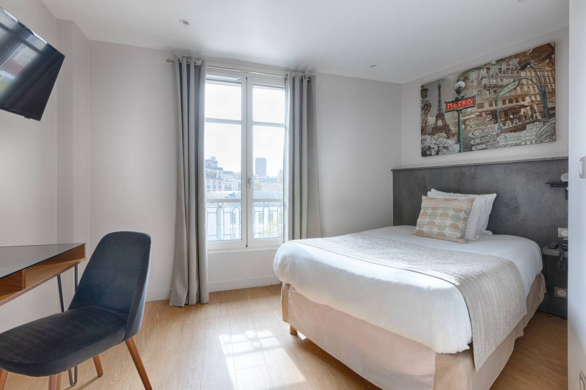 A hotel room with a bed, a desk, a chair, a window with curtains, and a cityscape view.