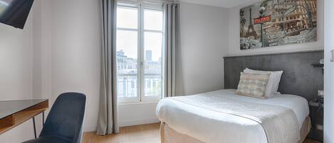 A hotel room with a bed, a desk, a chair, a window with curtains, and a cityscape view.
