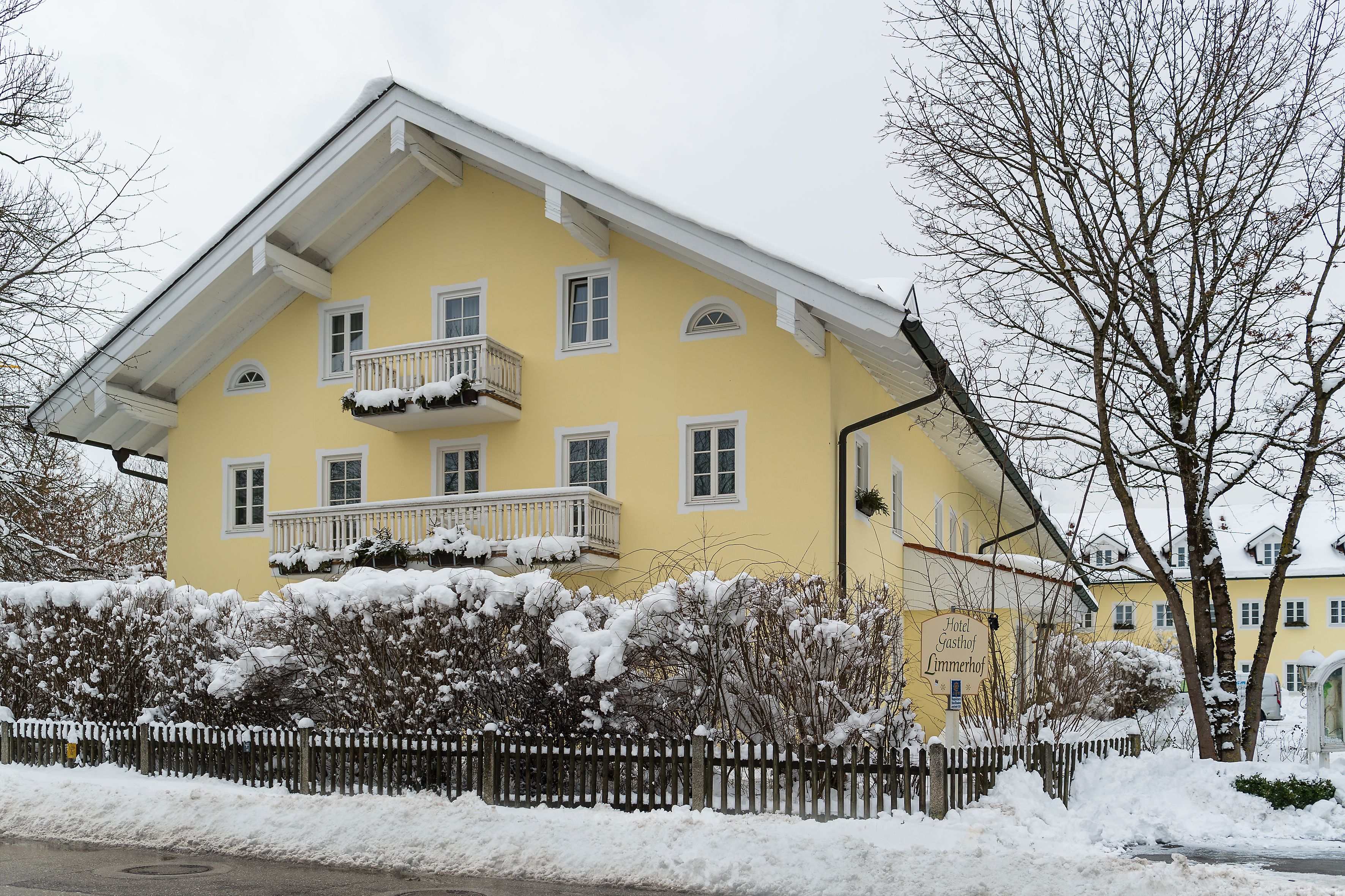 A yellow building with a balcony, surrounded by snow-covered bushes and a wooden fence.