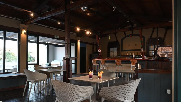An indoor dining area with high ceilings, wooden beams, and large windows. Tables and chairs are arranged for dining. - Sungate (Corfu)