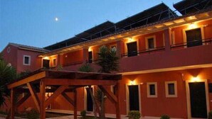 A two-story building with a red facade, multiple windows, and a covered outdoor area with a wooden structure. - Sungate (Corfu)