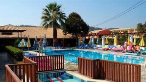 A swimming pool with lounge chairs and umbrellas, surrounded by wooden fences and palm trees. - Sungate (Corfu)