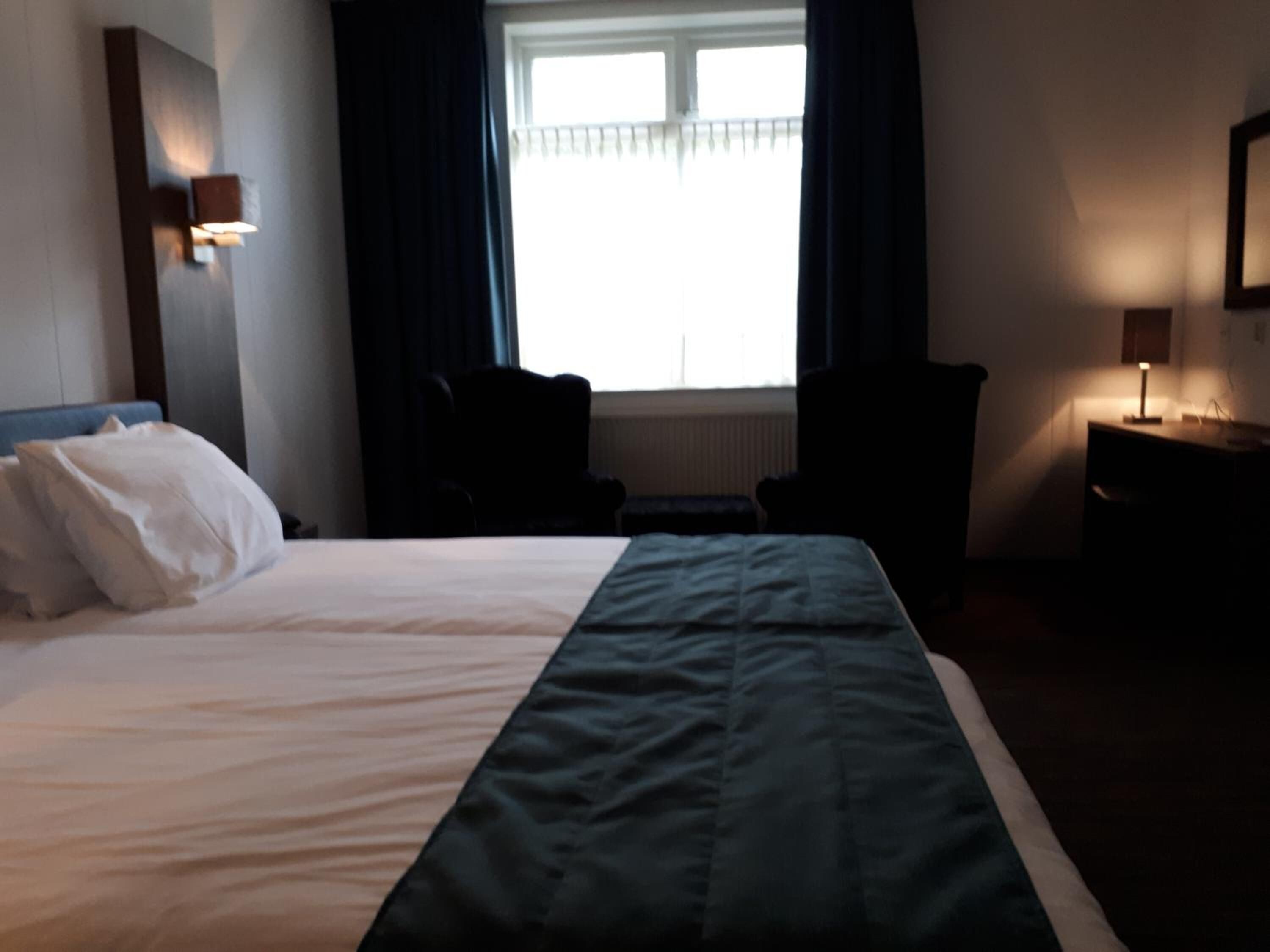 Una habitación de hotel con cama, escritorio, silla y ventana con cortinas.