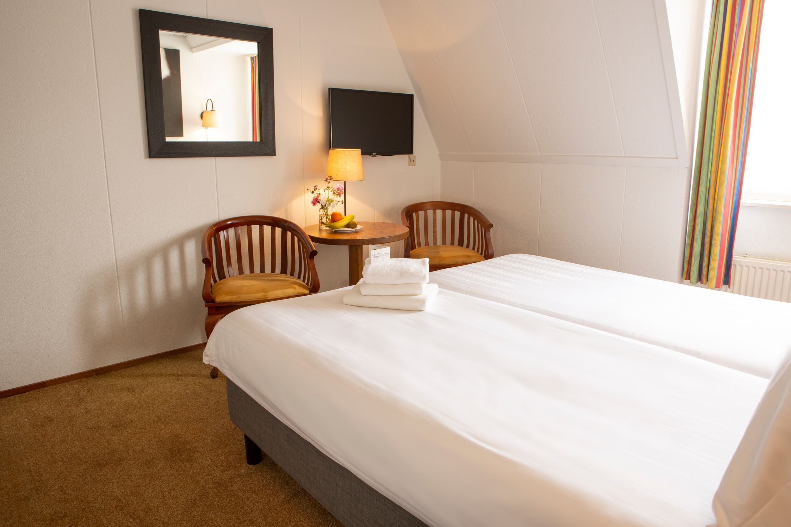 Ein Hotelzimmer mit einem großen Bett, zwei Holzstühlen, einem kleinen Tisch mit einer Lampe, einem Flachbildfernseher und einem Fenster mit Vorhängen.