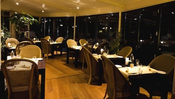 Restaurante com piso de madeira aconchegante e cadeiras de vime. Mesas montadas com velas e copos.