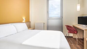 Une chambre d’hôtel comprenant un lit, un bureau avec une chaise, une télévision, une fenêtre et un luminaire fixé au mur.