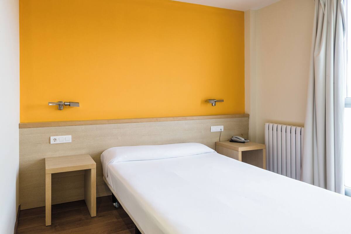 A hotel room with a single bed, two bedside tables, a nightstand with a phone, and a window with curtains.