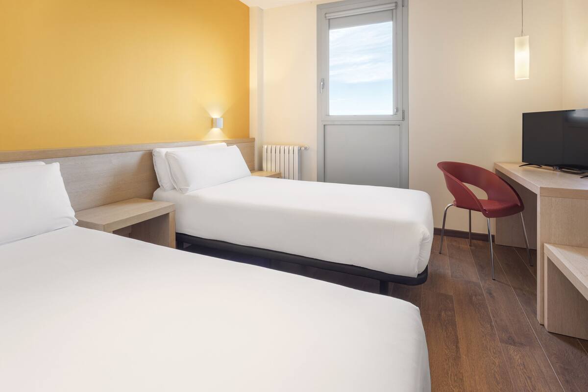 A hotel room with two beds, a desk with a chair, a television, and a window.