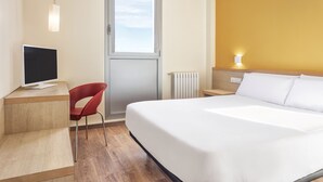 Habitación de hotel con cama, escritorio con computadora, una silla roja, ventana y mesita de noche con lámpara.