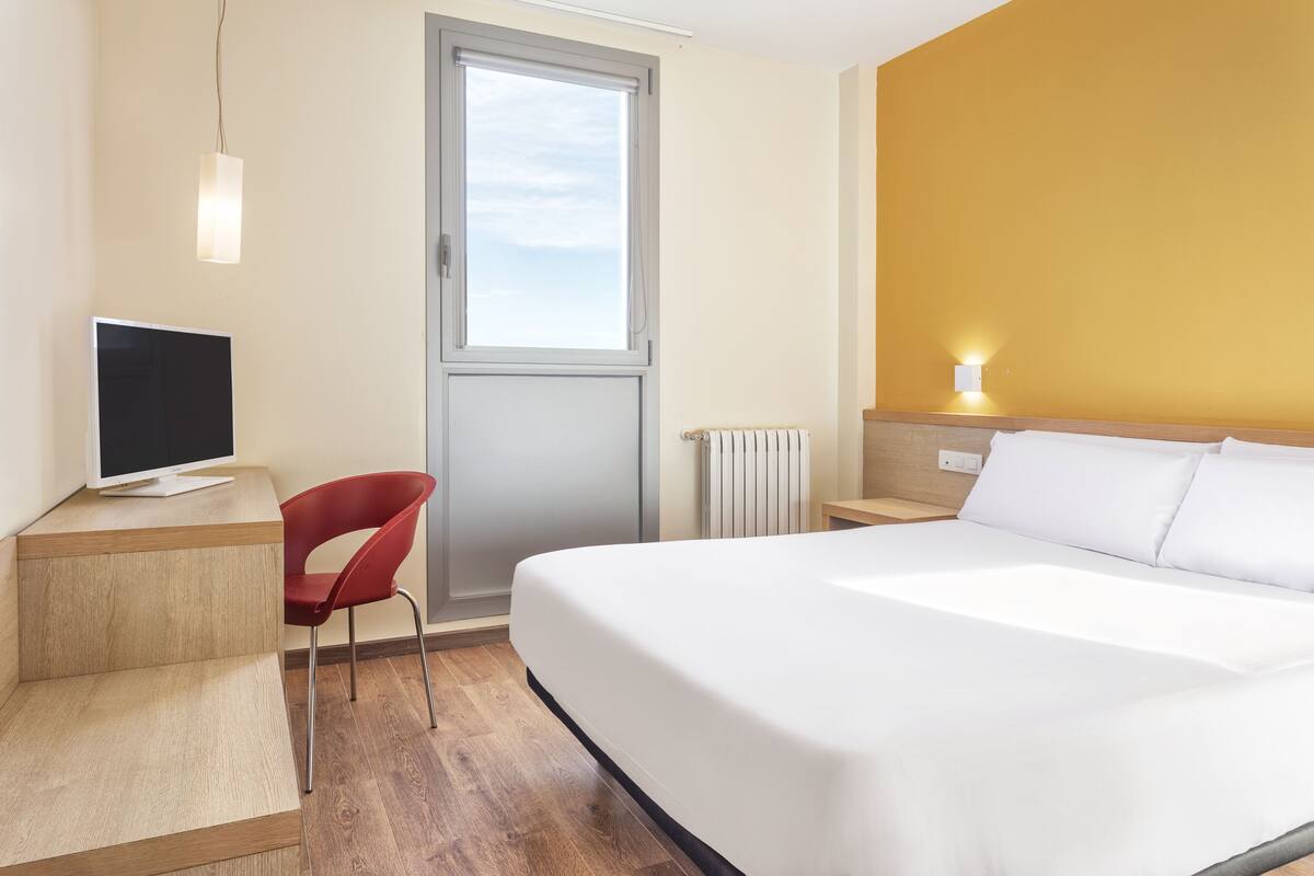 A hotel room with a bed, a desk with a computer, a red chair, a window, and a bedside table with a lamp.