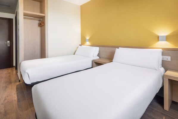 Habitación de hotel con dos camas individuales, cabeceros de madera, un armario de madera y una lámpara fijada a la pared.