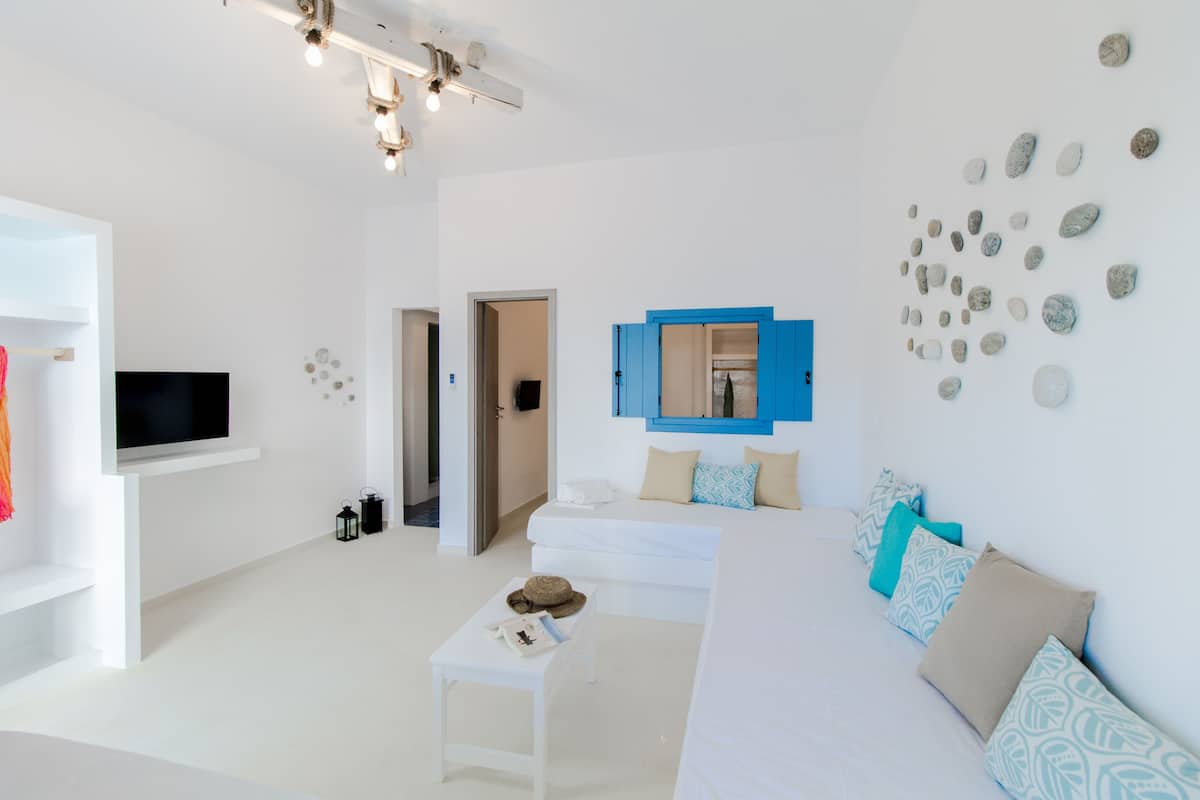A modern, minimalist bedroom with a white color scheme, a flat-screen TV, a bed with patterned pillows, a small table with a hat and book, and a wall with decorative stones.