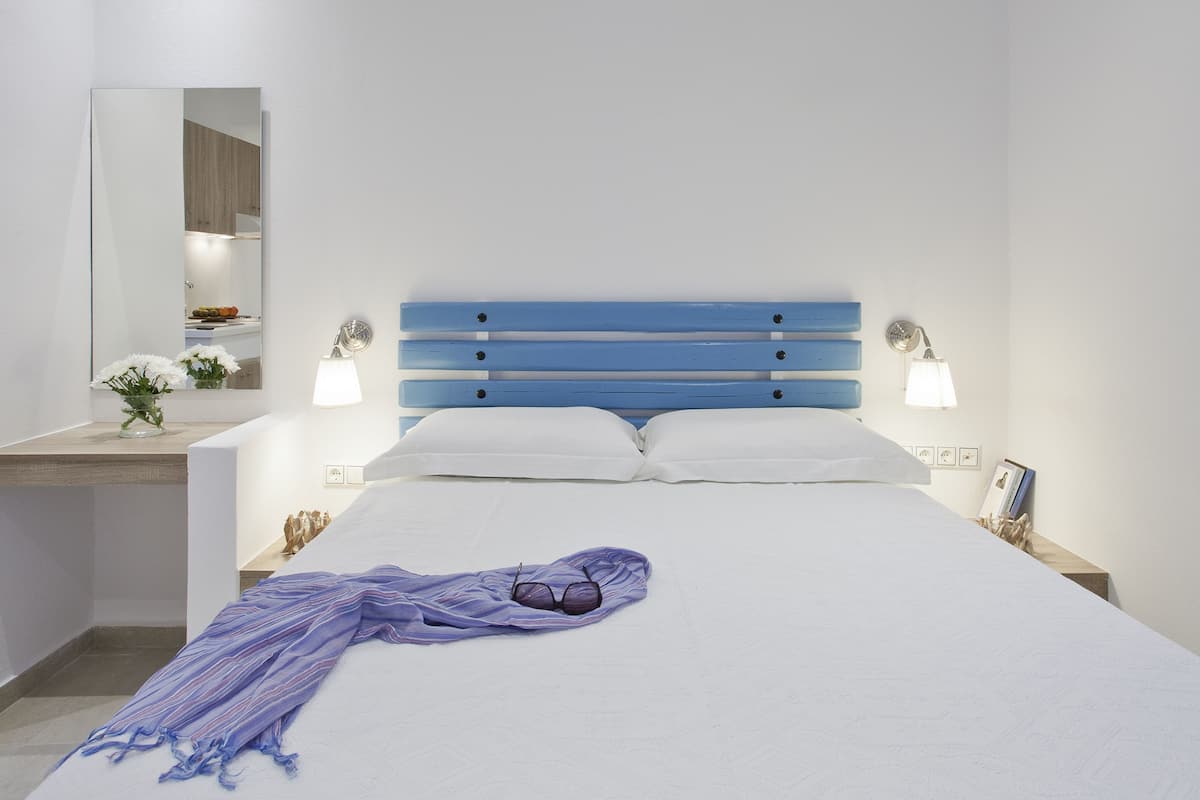 A neatly made bed with a blue headboard, white pillows, and a purple blanket. There is a bedside table with a lamp and a small plant.