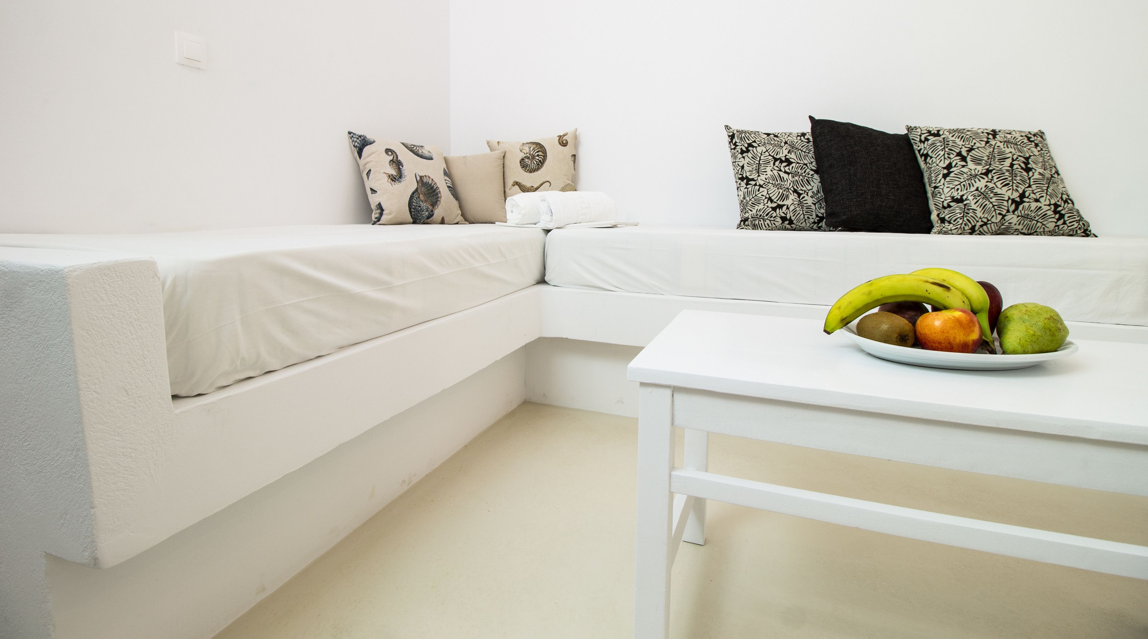 A white sofa with a white cushion, a white table with a fruit bowl, and patterned pillows on the wall.