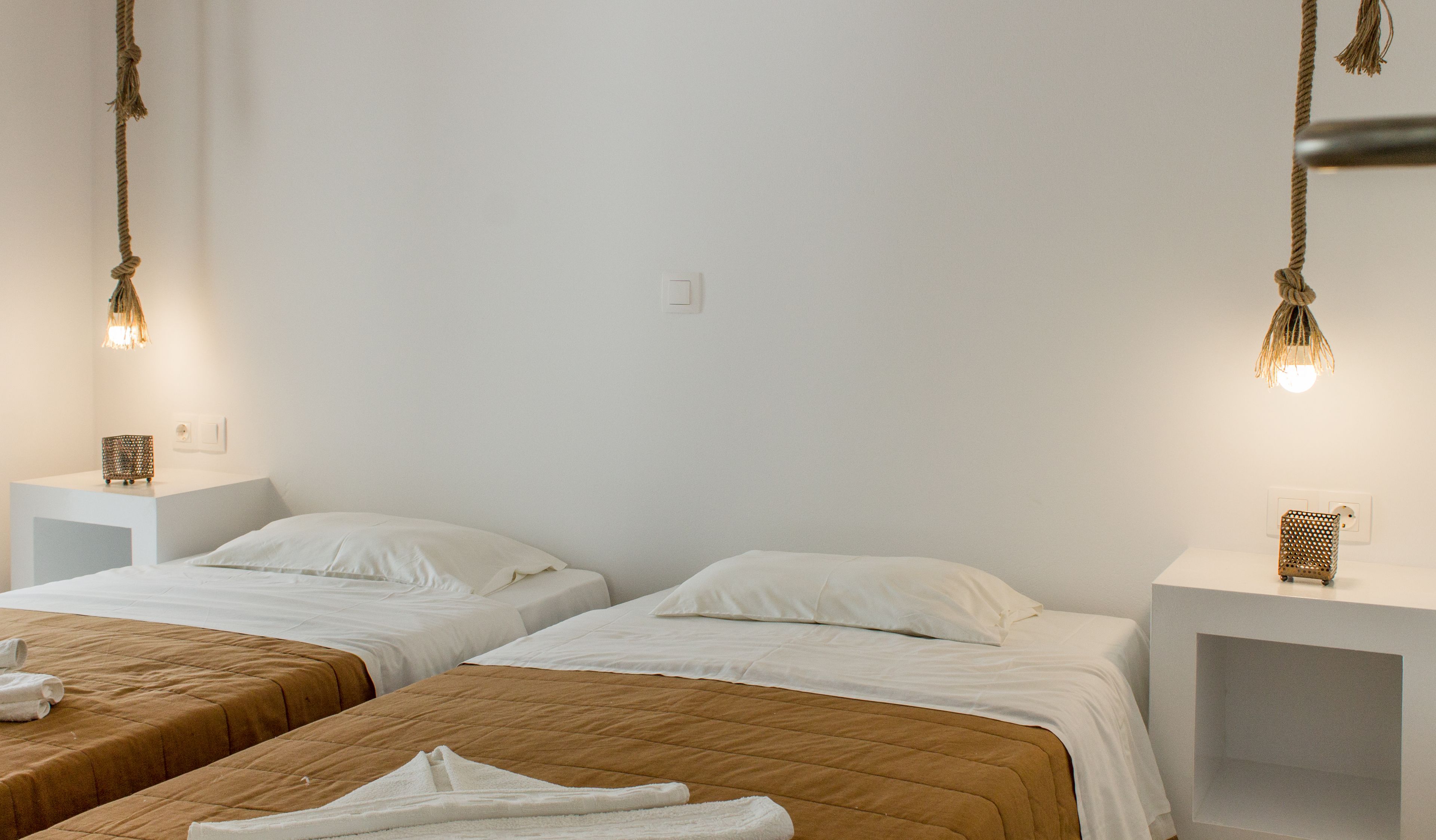 A hotel room with two beds, each with a white pillow and a brown bedspread. There are bedside tables with lamps and a small decorative item on each.