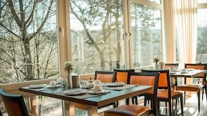 Een eetruimte met tafels voor vier personen, grote ramen en uitzicht op bomen.
