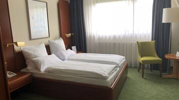 A hotel room with two single beds, a wooden nightstand with a lamp, a chair, and a small table with a lamp.